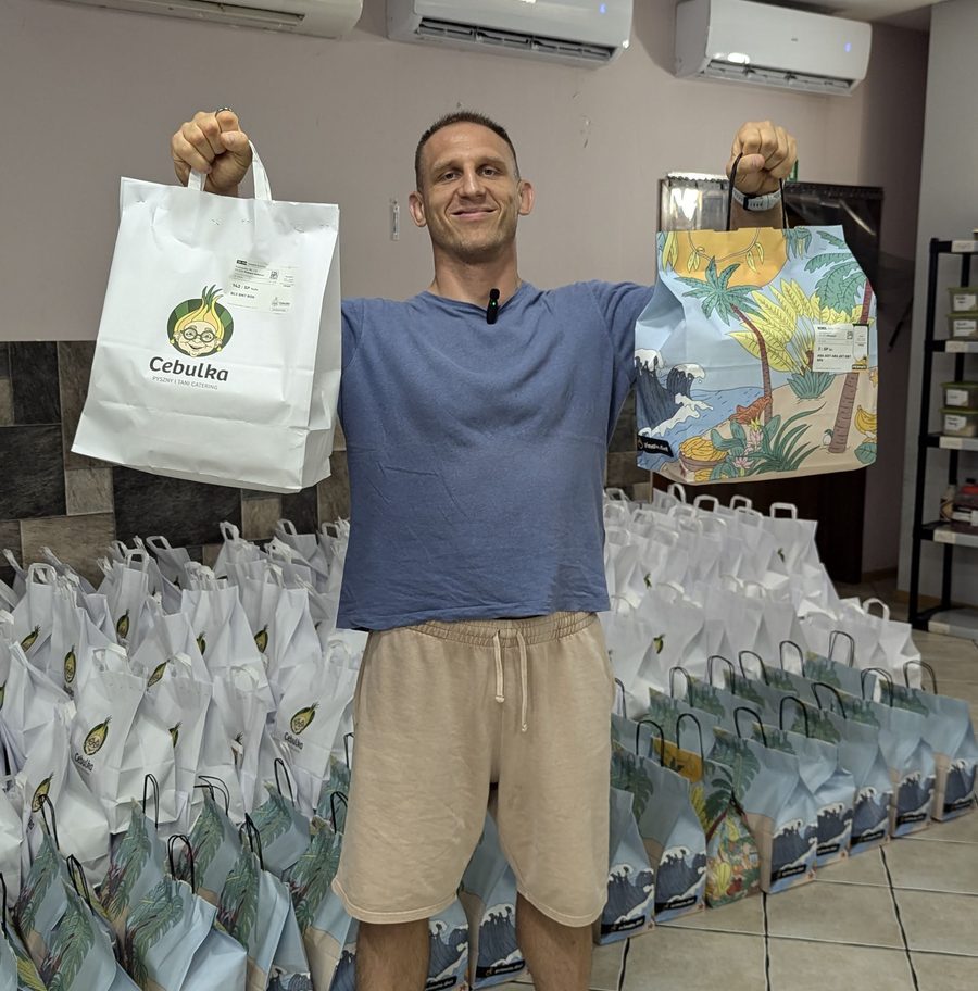 Paweł Kaczyński holding two branded meal delivery bags in front of rows of packed bags ready for the morning run
